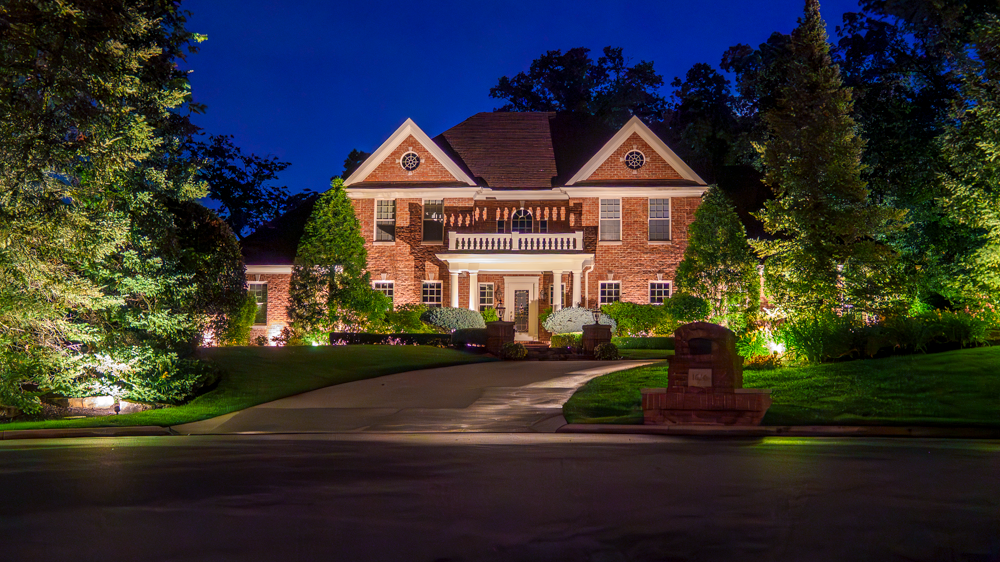 Warm lighting across a brick home, front landscaping and mature trees at dusk