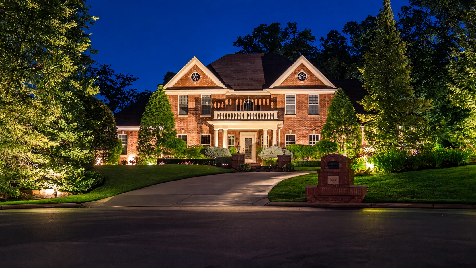 Warm lighting across a brick home, front landscaping and mature trees at dusk